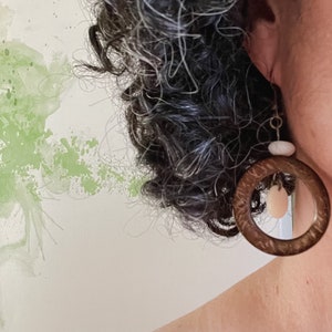 May include: A close-up of a brown hoop earring with a light-colored stone and a smaller, oval-shaped stone hanging inside. The earring is worn on a person's ear, with their curly hair and shoulder visible in the frame.