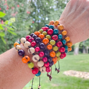 May include: A stack of colorful bracelets made from natural seed beads. The bracelets are tied with a black cord and feature a variety of colors, including red, orange, yellow, green, blue, and purple.