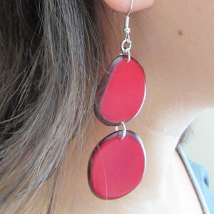 May include: A pair of red and black dangle earrings with a silver hook. The earrings are made of two round pieces of wood, one larger than the other, with a red finish. The earrings are hanging from a silver hook.