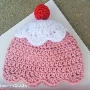Puede incluir: Un gorro de ganchillo rosa y blanco con forma de cupcake con una cereza roja de ganchillo en la parte superior.