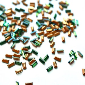 May include: A collection of small, rectangular, faceted beads in alternating metallic copper and deep emerald green colors. Each bead features a flat top surface with angled sides, scattered across a plain white surface in a loose arrangement.