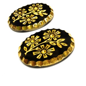 May include: Two oval-shaped buttons featuring a black base with a gold-colored floral design. Each button has a scalloped gold-colored edge and displays a raised pattern of two flowers on a single stem with leaves centered on the black surface.