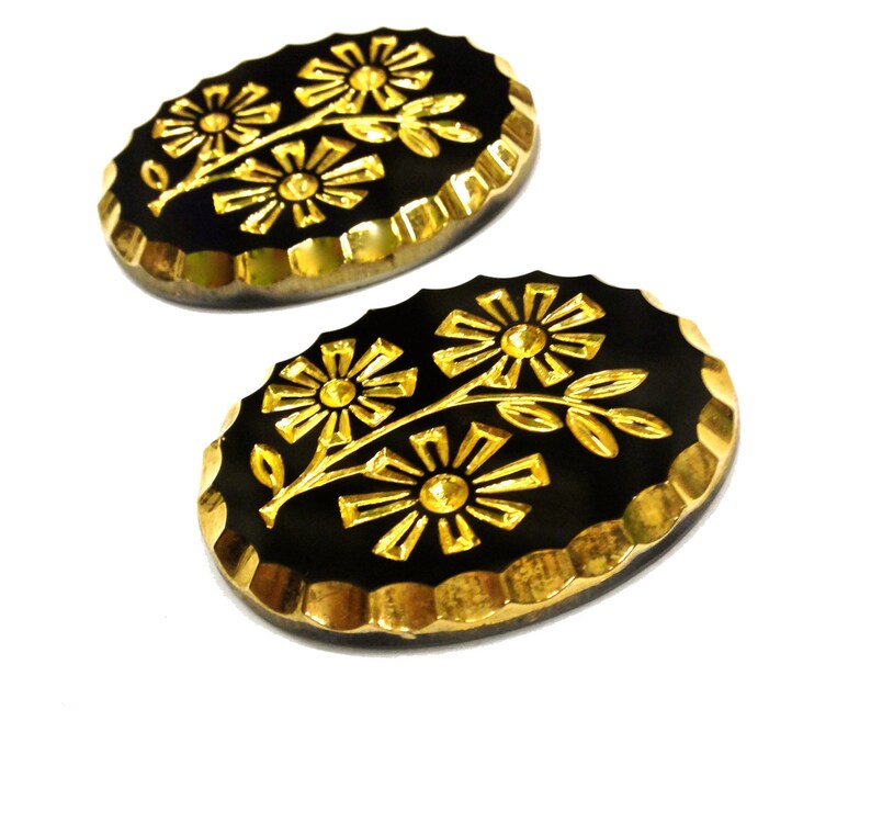 May include: Two oval-shaped buttons featuring a black base with a gold-colored floral design. Each button has a scalloped gold-colored edge and displays a raised pattern of two flowers on a single stem with leaves centered on the black surface.
