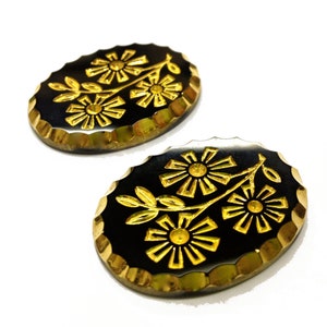 May include: Two oval-shaped buttons with a black base and a gold-colored floral design featuring three daisies on a stem. The buttons have a scalloped, metallic gold edge and a smooth, reflective surface finish on the black center.