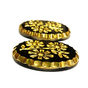 May include: Two oval-shaped buttons featuring a black center with a raised gold floral design. The edges of the buttons are finished with a metallic gold scalloped border, providing a distinct contrast against the dark, smooth central surface.