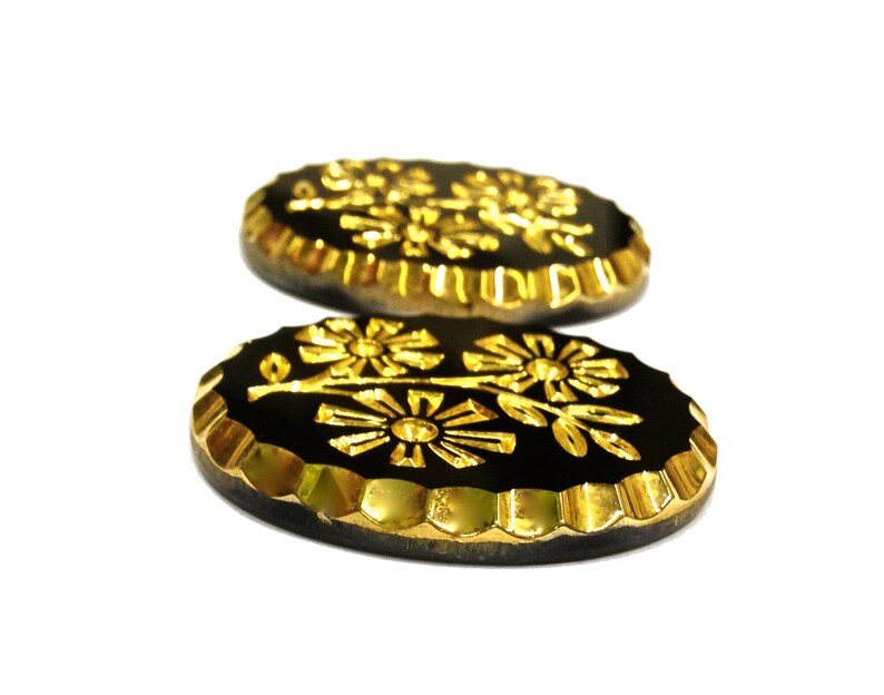 May include: Two oval-shaped buttons featuring a black center with a raised gold floral design. The edges of the buttons are finished with a metallic gold scalloped border, providing a distinct contrast against the dark, smooth central surface.