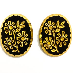 May include: A pair of oval-shaped earrings featuring a black background with a raised gold-tone floral design. Each earring has a scalloped gold edge and depicts three stylized flowers on a single stem with leaves, creating a high-contrast look.