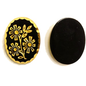 May include: Two oval-shaped buttons with a black surface. One button features a gold-colored embossed floral design with three flowers and leaves, surrounded by a scalloped gold edge. The second button is a plain, solid black oval with a smooth finish.