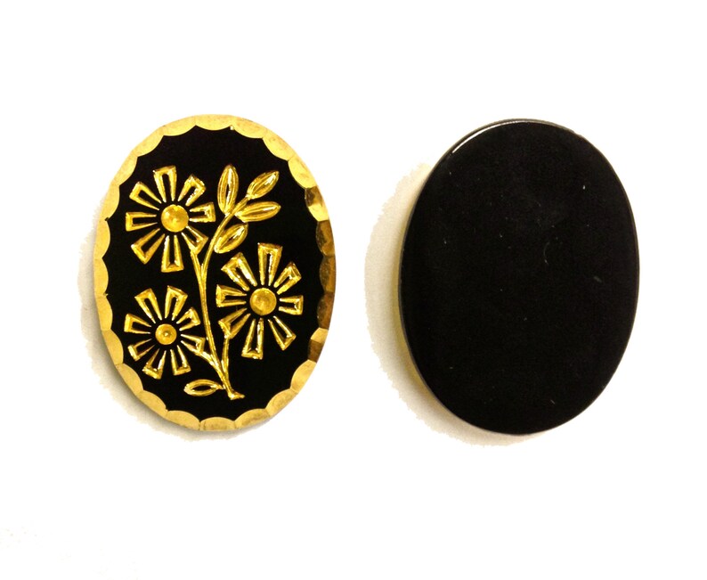 May include: Two oval-shaped buttons with a black surface. One button features a gold-colored embossed floral design with three flowers and leaves, surrounded by a scalloped gold edge. The second button is a plain, solid black oval with a smooth finish.