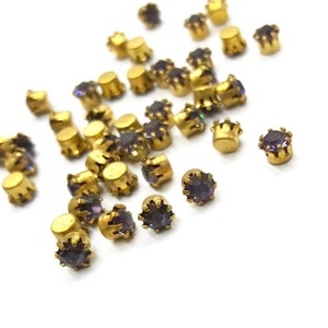 Puede incluir: Una colección de pequeñas piedras redondas de color púrpura facetadas, engastadas en monturas de metal dorado con garras. Cada pieza individual presenta una gema circular púrpura asegurada por múltiples garras metálicas, dispersas por el encuadre.