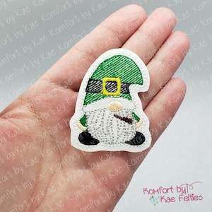 May include: Embroidered gnome patch with a green plaid hat, white beard, and black shoes. The gnome has a gold buckle and is holding a brown object. The patch is a feltie and has the text "Komfort by Kae Felties".