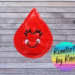 May include: A red felt drop-shaped applique with a smiling face and stitched details. The applique has black eyelashes and a white mouth. A circular logo with the text "Komfort by Kae FELTIES" is in the lower right corner.