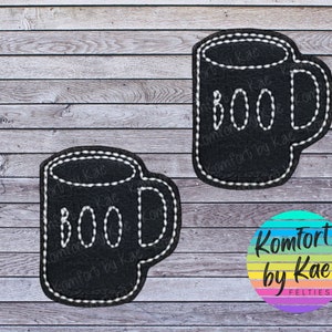May include: Two black felt mug-shaped appliques with white stitched outlines. Each mug has the word "BOO" stitched in white. The appliques are on a wooden background. The bottom right corner has a colorful logo that says "Komfort by Kae Felties".
