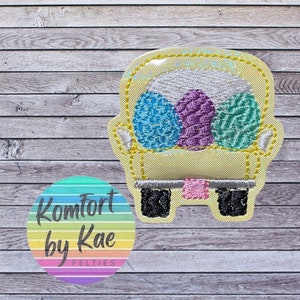 May include: A yellow felt applique of a couch with three balls of yarn inside. The couch has a pink and white stripe on the bottom. The applique is on a wood background with a circle that says "Komfort by Kae FELTIES".