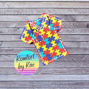 May include: A colorful felt item shaped like a shirt, featuring a puzzle piece design in red, yellow, blue, and green. The item is stitched with white thread. A circular logo with the text "Komfort by Kae FELTIES" is in the lower left corner.