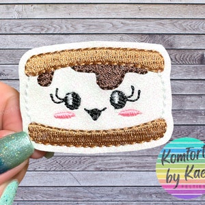 May include: A white felt applique of a smiling s'more with brown chocolate and graham cracker details. The s'more has pink cheeks and black eyelashes.