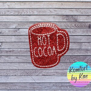 May include: A red glittery felt mug with the words "HOT COCOA" in white stitching. The mug has a white stitched outline and a handle. The background is a gray wooden surface.