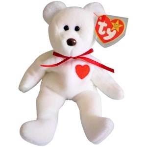 Può includere: Orso di peluche bianco Beanie Baby con un cuore rosso sul petto e un nastro rosso intorno al collo. L'orso ha occhi neri e un naso marrone. L'etichetta dice "Beanie Babies Baby Ty".