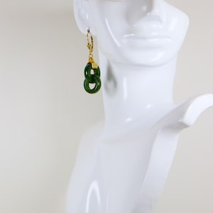May include: A pair of gold-tone earrings with a green jade design. The earrings feature a lever back closure and two interlocking green jade rings. The earrings are displayed on a white mannequin head.