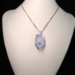 Puede incluir: Un collar con colgante de jade tallado. El colgante es de color morado claro y verde, con intrincadas tallas. El collar tiene una delicada cadena dorada. El colgante se exhibe en un maniquí blanco.