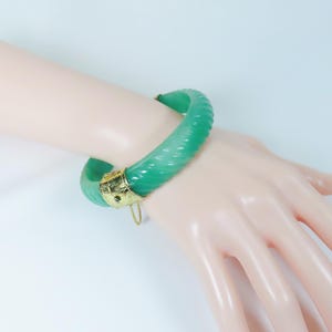 May include: A jade green bangle bracelet with a textured surface and gold-colored accents. The bracelet is worn on a wrist, showcasing its color and design. The bangle is a piece of jewelry.