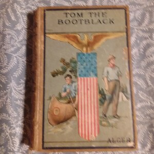 May include: A vintage book cover with the title "Tom the Bootblack" in bold, black lettering. The cover features a patriotic illustration of an eagle with outstretched wings holding an American flag. Two boys are depicted in the foreground, one paddling a canoe and the other standing beside him. The book cover is in a worn condition with a faded blue background.