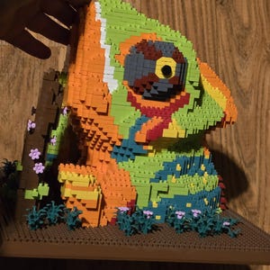 May include: A colorful chameleon sculpture made of interlocking plastic bricks. The sculpture features shades of green, orange, blue, and brown, with a detailed eye and a small tree. The sculpture is displayed on a brown base with small plants and flowers.
