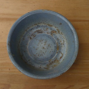May include: A round, light blue metal bowl with a slightly textured surface. The bowl shows signs of age with areas of rust and wear. The bowl is sitting on a wooden surface.