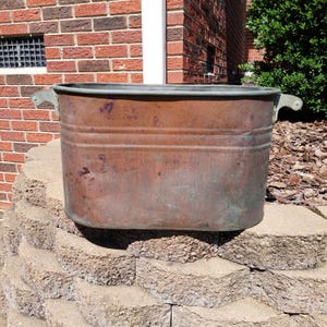 May include: An oval-shaped copper wash tub featuring a weathered, oxidized patina with visible dark spots and surface wear. The vessel includes two riveted metal handles on opposite sides and two horizontal embossed ridges encircling the body.
