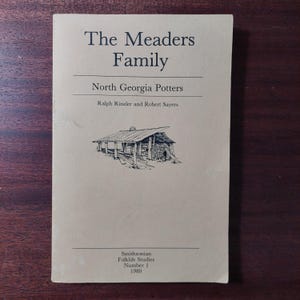 May include: A vintage book titled "The Meaders Family" with the subtitle "North Georgia Potters." The cover is a light tan color with black text and a black and white illustration of a rustic building. Published by Smithsonian Folklife Studies in 1980.