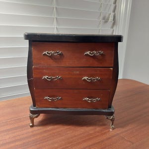 May include: A small, antique wooden chest of drawers with three drawers. The chest has a dark finish on the top and base, with a reddish-brown finish on the drawers. Each drawer has ornate metal handles. The chest sits on decorative metal feet.