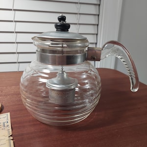 May include: A clear glass teapot with a silver band and a clear glass handle. The teapot has a built-in strainer for loose tea leaves. The lid is silver with a black knob.