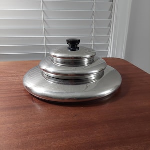May include: A set of three stainless steel nesting bowls with a black knob on the lid. The bowls are stacked on a wooden surface, with the largest bowl at the bottom and the smallest on top.
