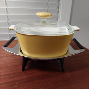 May include: A vintage yellow casserole dish with a clear glass lid and a yellow knob. The dish sits on a metal warming stand with black handles. A classic kitchenware item.