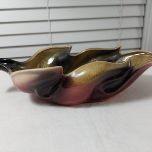 May include: A ceramic leaf-shaped bowl with a red, brown, and green glaze. The bowl has a unique, organic shape and a handle on one side.