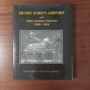May include: A black and white photo of a book cover titled "Henry Ford's Airport and Other Aviation Interests 1909-1954" by Timothy J. O'Callaghan. The cover features a black and white photo of a building with a sign that reads "Ford Airport".