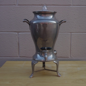 May include: A vintage silver-toned coffee percolator with a clear glass lid and black spigot. The coffee maker has two curved handles and sits on three decorative legs. The percolator is displayed on a light brown wooden surface.