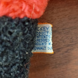 May include: Close-up of a blue and orange fabric tag with the text "DISNEY PRODUCTS CONTENT ALL NEW FIBERS TREATED AROUND N.Y. WALT DISNEY", attached to a red and blue plush toy.