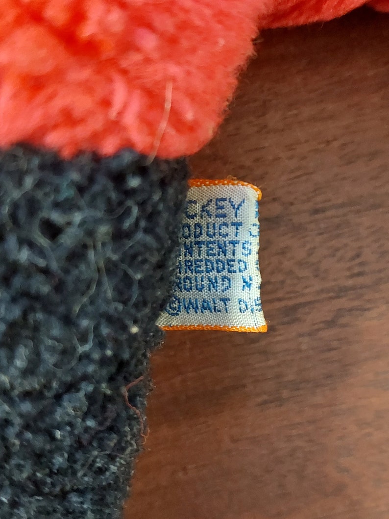 May include: Close-up of a blue and orange fabric tag with the text "DISNEY PRODUCTS CONTENT ALL NEW FIBERS TREATED AROUND N.Y. WALT DISNEY", attached to a red and blue plush toy.