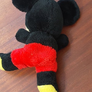 May include: Black and red plush Mickey Mouse toy with yellow accents. The toy is lying on its back with its legs in the air.