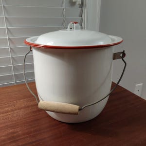 May include: A white enamel bucket with a matching lid, featuring a thin red rim around the top edge and lid. The bucket includes a metal wire handle with a light-colored wooden grip, attached to metal brackets on the sides of the cylindrical body.