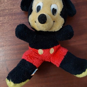 May include: A black and red plush Mickey Mouse toy with large eyes and a big smile. The toy is wearing red shorts with two yellow buttons.