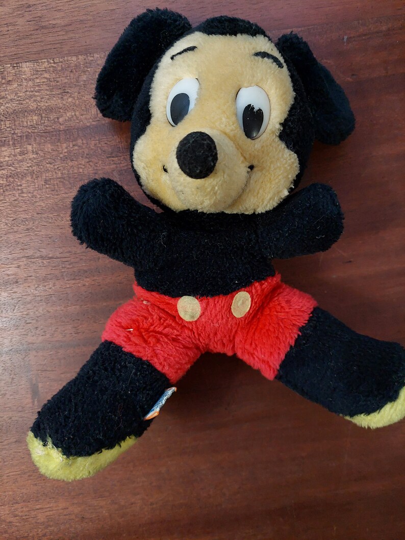 May include: A black and red plush Mickey Mouse toy with large eyes and a big smile. The toy is wearing red shorts with two yellow buttons.