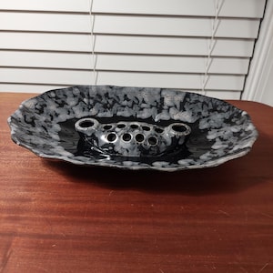 May include: A black and white ceramic flower frog with a speckled pattern. The frog has a series of holes for arranging flowers and is set in a matching oval dish. The dish has a wavy edge.