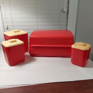 May include: A set of four red and cream colored kitchen canisters. The set includes a bread box and three smaller canisters.
