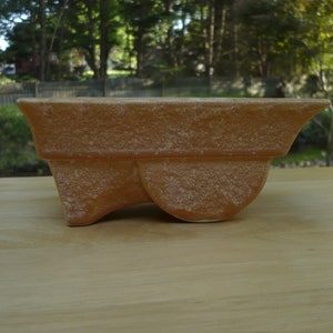 May include: A rectangular, light brown planter with a textured surface. The planter has a unique, curved design with a rounded base and flared sides. The planter is made of ceramic and is suitable for indoor or outdoor use.