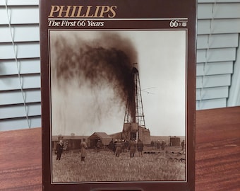 May include: A brown hardcover book titled Phillips The First 66 Years. The cover features a sepia-toned historical photograph of an oil derrick emitting dark smoke, with several people standing in a field in the foreground near small buildings.