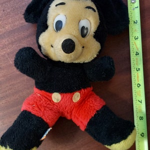 May include: A black and yellow plush Mickey Mouse toy with red shorts and black button eyes. The toy is about 25 cm tall and is sitting on a wooden surface.