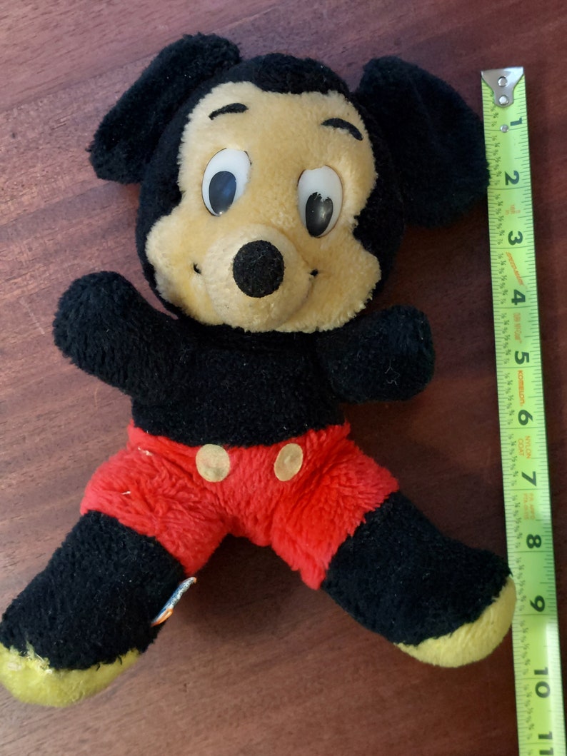 May include: A black and yellow plush Mickey Mouse toy with red shorts and black button eyes. The toy is about 25 cm tall and is sitting on a wooden surface.