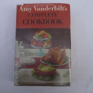 May include: A red hardcover cookbook with the title "Amy Vanderbilt's Complete Cookbook" on the front cover. The cover features a photo of a roasted turkey, a bowl of salad, and a fruit bowl with apples, grapes, and bananas.